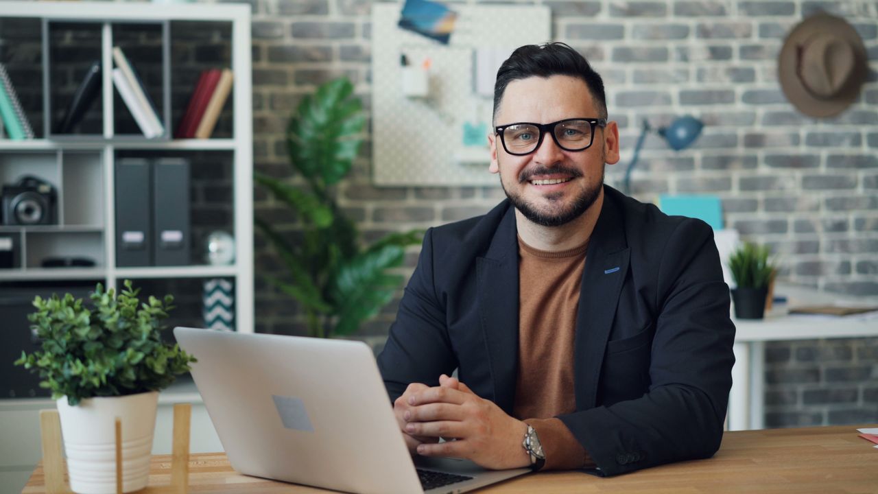 Businessman with eyeglasses smiling while working on a laptop in a modern office.